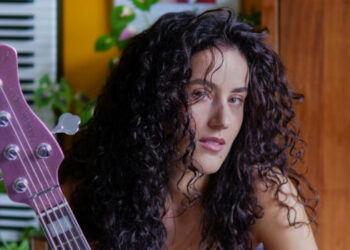 Nadia Essah with curly hair holding a guitar in a colourful room.