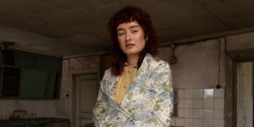 Woman with curly hair in vintage setting, wearing floral blazer and yellow blouse.